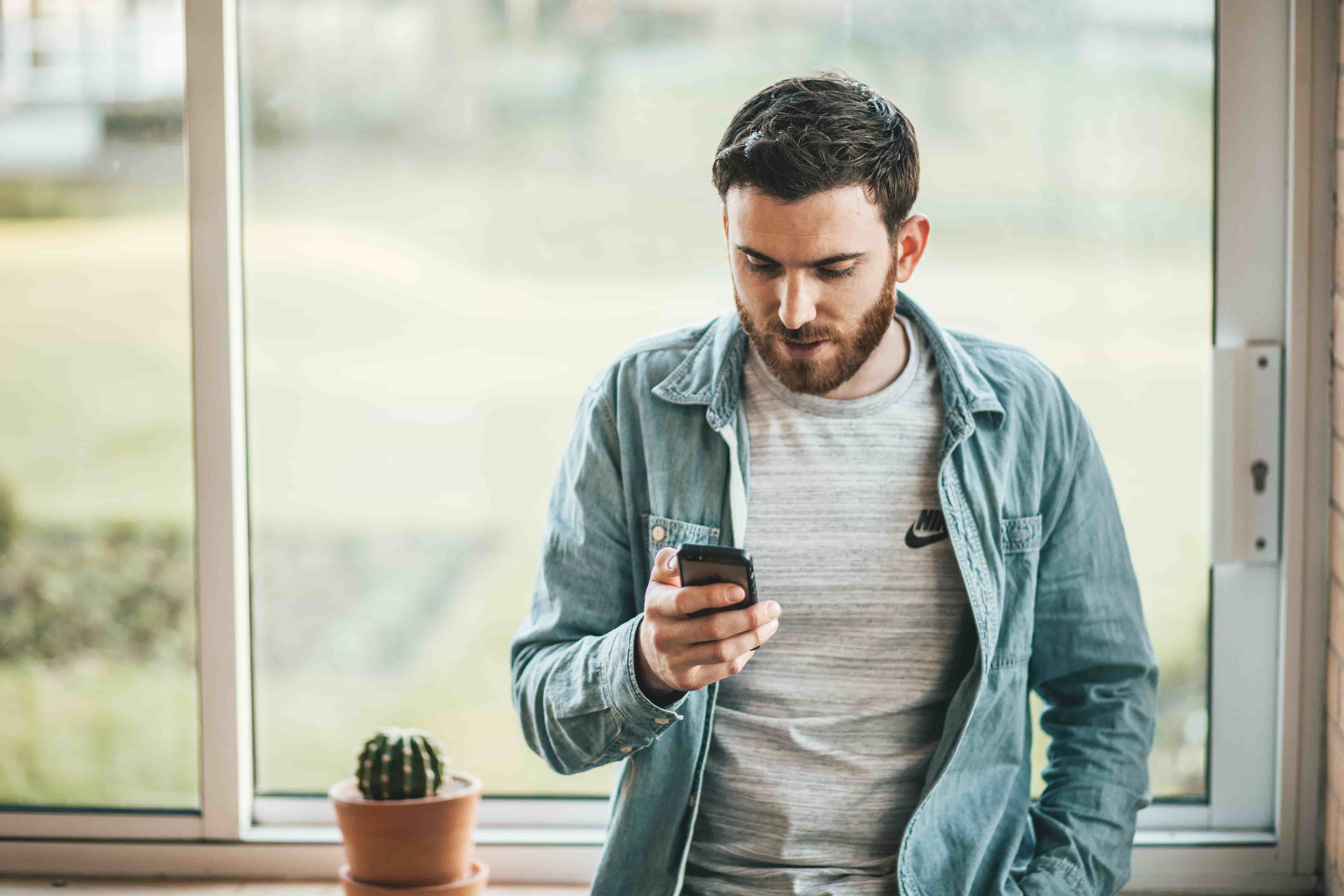 bearded man in casual clothing leans against windowsill looking at his cell phone to track his net worth